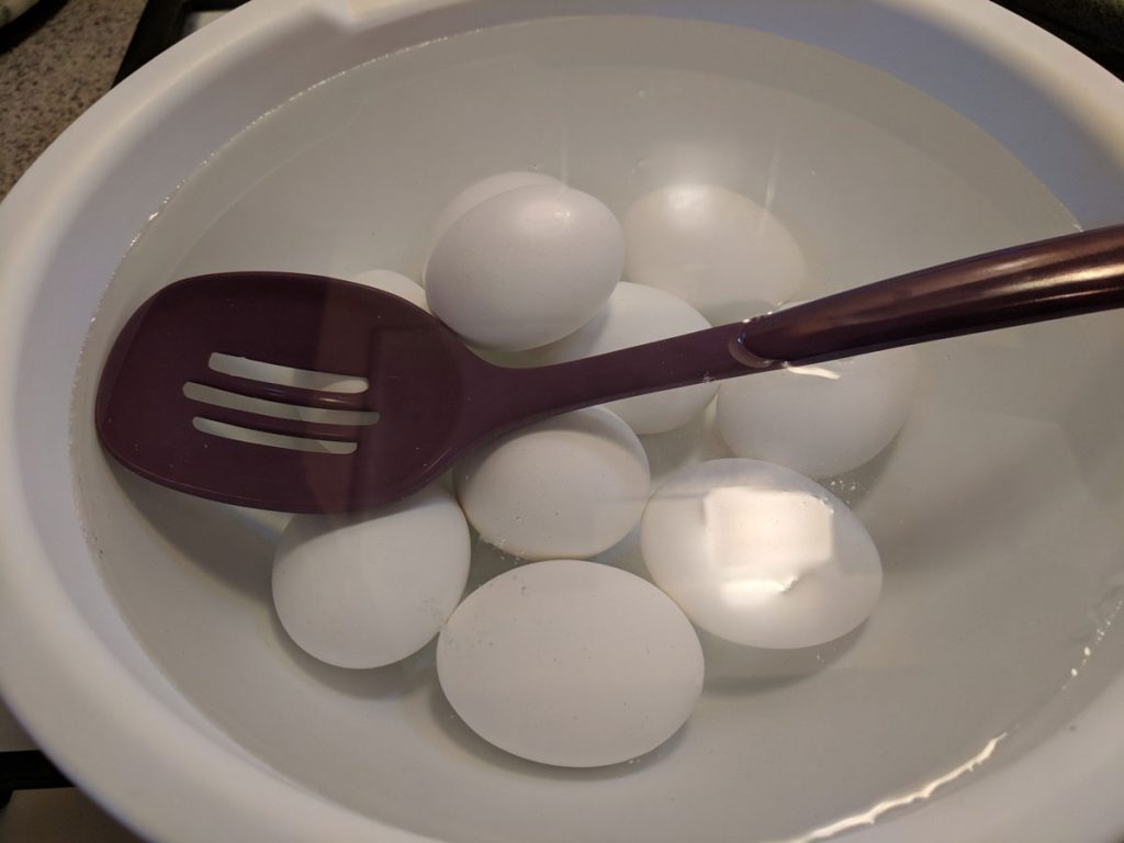 A white bowl full of water and eggs, with a purple slotted spoon 