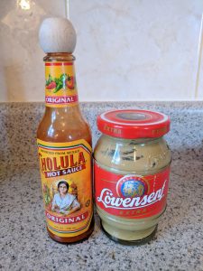 A bottle of Cholula hot sauce next to a jar of Lowensenf Extra mustard on a speckled kitchen counter